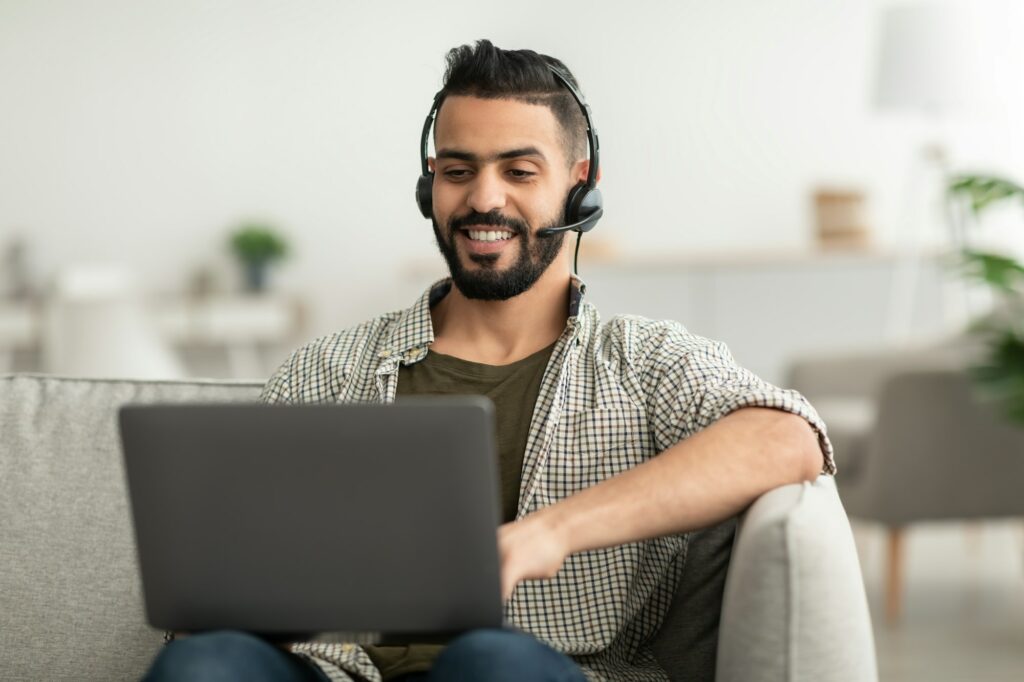 Young happy Arab man in headphones using laptop pc, working online from home